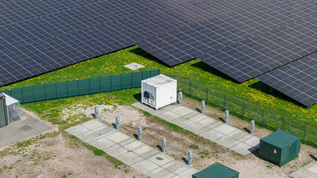 Aerial view of a solar panel farm featuring rows of photovoltaic modules, a white utility container, and a green fence on a grassy field in Warmenhuizen, North Holland, Netherlands.