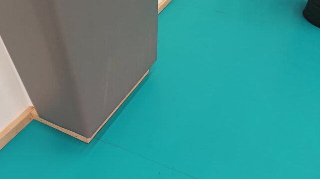 Gray cabinet corner positioned against bright turquoise floor with wooden trim, showcasing gradual movement and spatial relationship in a modern interior setting