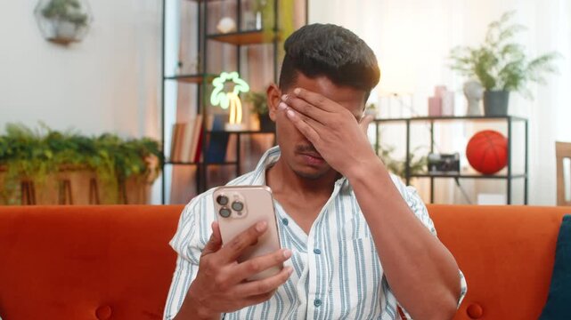 Indian man at home reads smartphone alert about game loss and sighs feels upset and disappointed. Arabian guy on sofa stares at screen after defeat rubs face breathes out and drops head with sad mood.