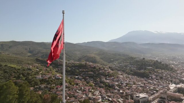 Aerial panoramic view Albanian national flag prominently wave atop stone fortification at Berat Castle - Kalaja e Beratit under clear blue sky, overlooking lush, forested hillside and Berat cityscape