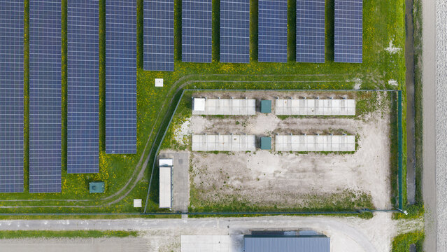 Aerial view of rows of solar panels on a green field with yellow wildflowers and utility infrastructure in Warmenhuizen, North Holland, Netherlands.