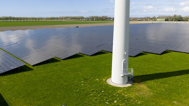 Aerial view of a large solar farm with rows of photovoltaic panels next to a wind turbine base in a green field under a clear sky Warmenhuizen, North Holland, Netherlands.