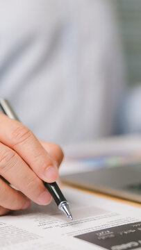 Detailed view showing male hand holding black pen writing signature paper document near credit card laptop computer inside bright office workspace professional business executive reviewing financial.