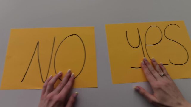 	
Close-up of a woman&rsquo;s hands sliding two paper cards labeled &ldquo;Yes&rdquo; and &ldquo;No&rdquo; across a table, finally leaving &ldquo;Yes&rdquo; in place. Minimal, clean visual illustrating decision-making, choice, 