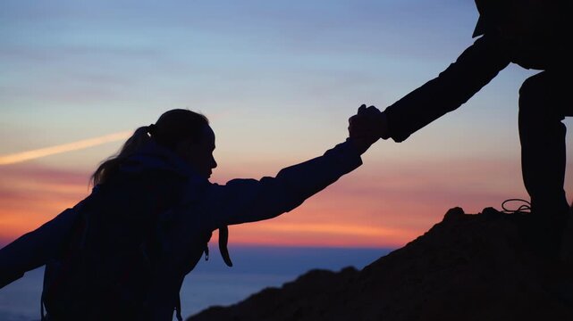 Couple of travelers climbing mountain at sunset, with man reaching out hand to help woman ascend steep rock. Concept teamwork, partnership and mutual support