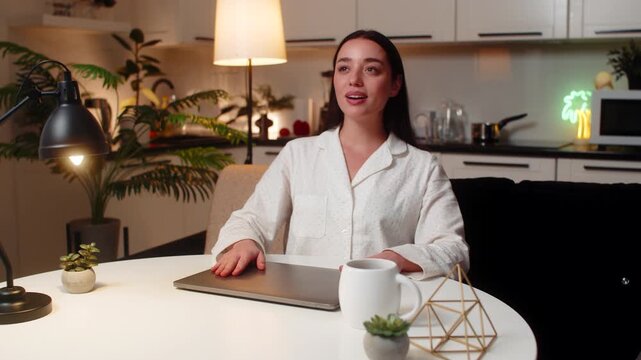 Young woman at night home ends tasks closes laptop exhales feels relief after long work. Caucasian girl on sofa at table at evening leans back rests shoulder calm breath enjoys pause feels calm mood.