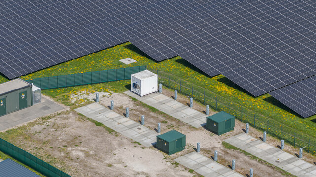 Aerial view of a large solar farm with rows of photovoltaic panels, technical equipment containers, and green grass with yellow flowers in Warmenhuizen, North Holland, Netherlands.