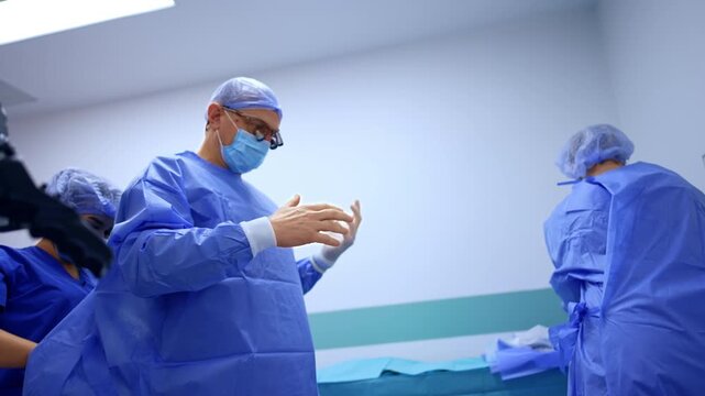 Male Caucasian doctor is helped to put on gloves and protective blue robe. Low angle view at the doctor preparing for neurosurgery.