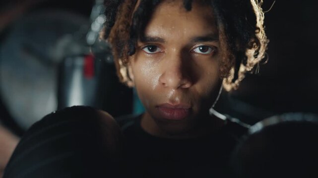 Attractive black man with boxing gloves looking at camera in dark gym, portrait. Closeup view of face of exhausted fighter after fight in box club, tired junior sportsman breathing hard and resting
