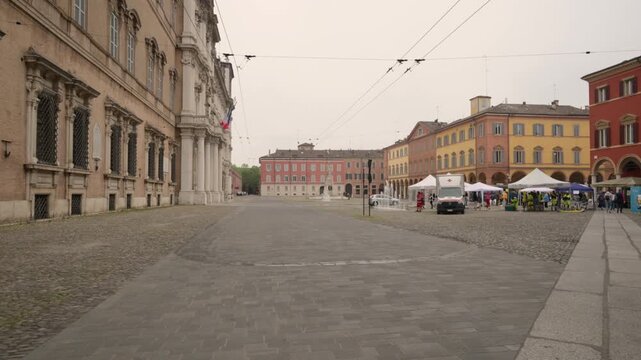 Modena, italy, april 12, 2026. First person perspective walking across the historic piazza roma in modena, italy, toward the ducal palace on a cloudy day
