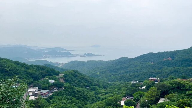 The panning video shows a view of the ancient village of Jiufen, Taiwan. Houses, streets, and lush green forests stand in the background, with mountains and the sea on a cloudy day with fog.