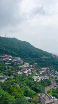 The panning video shows a view of the ancient village of Jiufen, Taiwan. Houses, streets, and lush green forests stand in the background, with mountains and the sea on a cloudy day with fog.