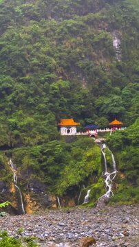 The vertical video shows a temple with a waterfall cascading down a green hillside in Taroko National Park, Taiwan. Lush mountain forests as a backdrop in the rain.