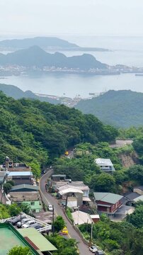 The vertical video shows a view of the ancient village of Jiufen, Taiwan. Houses, streets, and lush green forests stand in the background, with mountains and the sea on a cloudy day with fog.