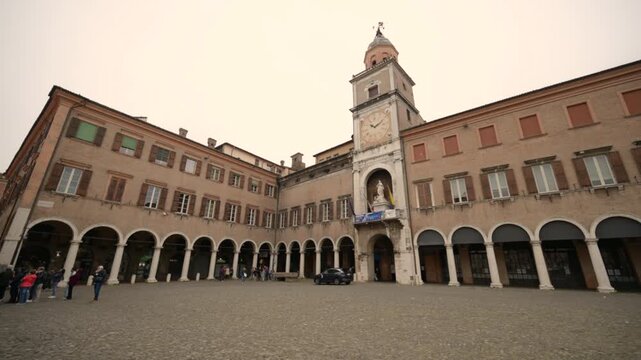 Modena, italy, april 12, 2026. Point of view walking through the cobblestone square of piazza grande in modena, italy. Historical building with arches and a clock tower
