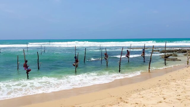 Ahangama, Sri Lanka - 26 March 2024: Horizontal video of fishermen fishing from wooden stilts in the middle of the sea off Sri Lanka. It's a sunny day with strong waves crashing against the shore.