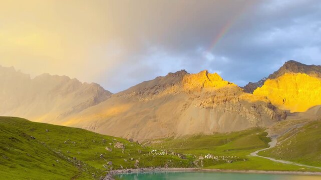 Video shows the front view of Tientang Hu Lake, Xinjiang, China. There are many mountains behind. The sun was setting with soft yellow light. There is a lush green lawn and a rainbow on a cloudy day.