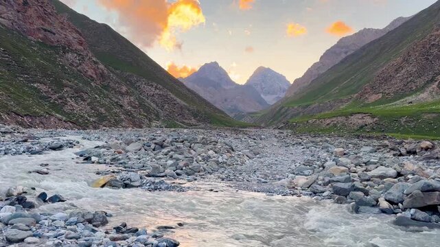 A wide-angle horizontal video shows the mountains, gently flowing rivers, and lush grassy hills of the Tian Shan Mountains in Xinjiang Province, China. Numerous rocks line the walking path at sunset.