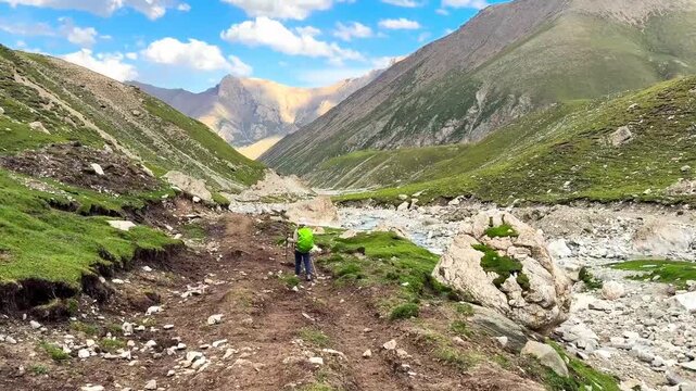 Horizontal video shows a hiker walking on green grasslands in the Tianshan Mountains in Xinjiang, China. There is a river flowing along the way. The idea for adventure activity footage.