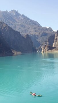 Panning video shows many boats sailing on Attabad Lake, Hunza, Pakistan. The backdrop features snow-capped mountains, poplar trees with yellow leaves, and the sparkling blue water on a sunny day.
