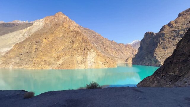 The video is from a boat sailing on Attabad Lake, Hunza, Pakistan. The backdrop features snow-capped mountains, poplar trees with yellow leaves, and the sparkling blue water on a sunny day.