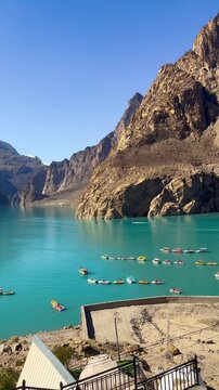 Panning video shows many boats sailing on Attabad Lake, Hunza, Pakistan. The backdrop features snow-capped mountains, poplar trees with yellow leaves, and the sparkling blue water on a sunny day.