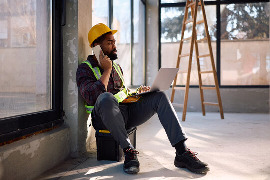 Black worker using laptop while talking on cell phone at construction site.