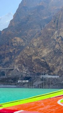The video is from a boat sailing on Attabad Lake, Hunza, Pakistan. The backdrop features snow-capped mountains, poplar trees with yellow leaves, and the sparkling blue water on a sunny day.