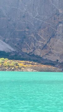 The video is from a boat sailing on Attabad Lake, Hunza, Pakistan. The backdrop features snow-capped mountains, poplar trees with yellow leaves, and the sparkling blue water on a sunny day.