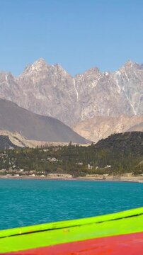 The video is from a boat sailing on Attabad Lake, Hunza, Pakistan. The backdrop features snow-capped mountains, poplar trees with yellow leaves, and the sparkling blue water on a sunny day.