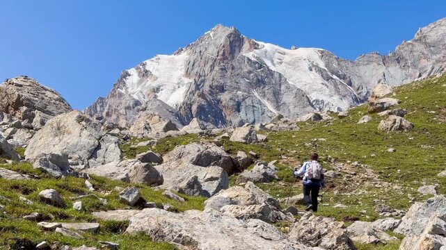 A wide-angle video shows a trekker walking on a grassy hill in the Trek Xinjiang route, China. The background is a mountain on a cloudy day. There are many rocks behind.