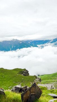 Panning video of a high-angle view of the snow-capped Himalayas and mist after rain at Bhrigu Lake Trek, Himachal Pradesh, India. There is a view of green hills layered and overlapping each other.