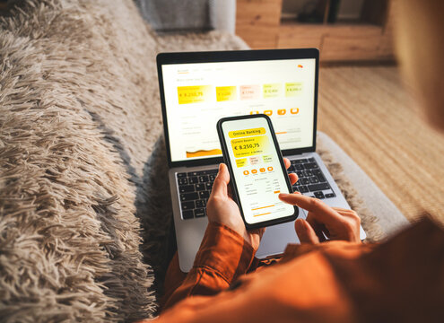 Woman using smartphone and laptop to manage personal budget while sitting on couch at home. Digital banking concept.