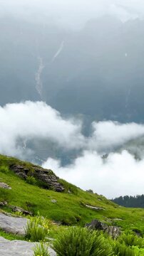 Panning video of a high-angle view of the snow-capped Himalayas and mist after rain at Bhrigu Lake Trek, Himachal Pradesh, India. There is a view of green hills layered and overlapping each other.