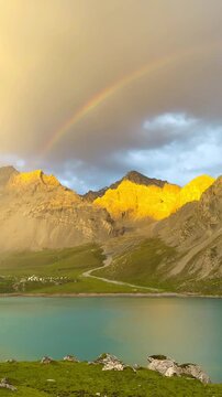 Video shows the front view of Tientang Hu Lake, Xinjiang, China. There are many mountains behind. The sun was setting with soft yellow light. There is a lush green lawn and a rainbow on a cloudy day.