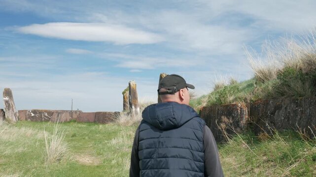 Handheld static shot of a man walking towards the ancient megaliths at the Great Salbyk Kurgan in Khakassia. A tourist explores the historical Valley of the Kings on a bright summer day.