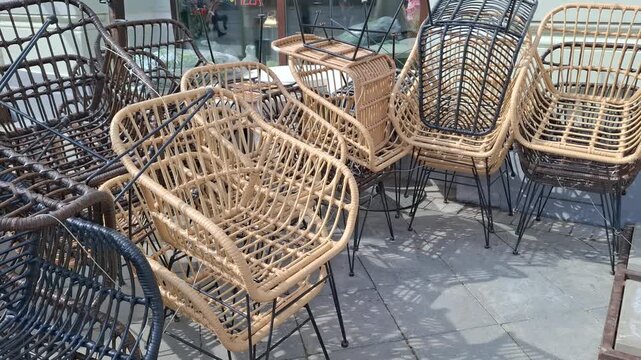 Collection of stylish wicker chairs stacked together outside cafe. Modern rattan seats in natural and dark tones. Elegant curved designs and thin black metal. For trendy patio look. Outdoor furniture