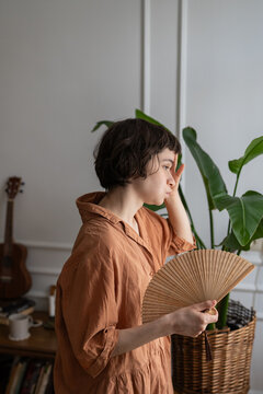 Woman suffering from overheating and headache standing in room waving hand fan for cooling, tired female annoyed by summer heatwave struggling with fatigue and migraine without air conditioning.