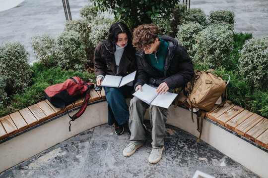 Two high school friends sit on a curved outdoor bench studying with open notebooks. They review notes together surrounded by backpacks and greenery.