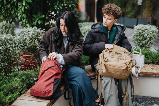 Two high school friends sit on a bench and check their backpacks. The casual outdoor scene shows teenagers talking and looking through their bags.