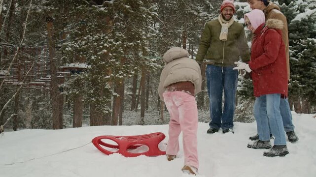 Group of four multiethnic gen Z friends playing rock paper scissors, African American girl beating them and sledding down hill, Caucasian man pushing her and she falling off sled