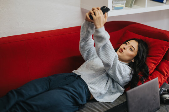 A female student lies on a red couch taking a selfie with her smartphone. She relaxes beside a laptop, blending study and leisure in a casual indoor setting.