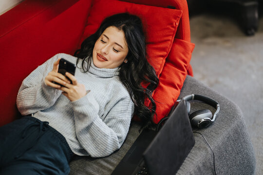 A young female student lies on a couch using her smartphone. She relaxes with a laptop and headphones nearby while scrolling on her phone.