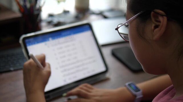 Over-the-shoulder view of a young Asian girl wearing glasses, focusing on her online lesson and using a stylus to write on a digital tablet at her home desk