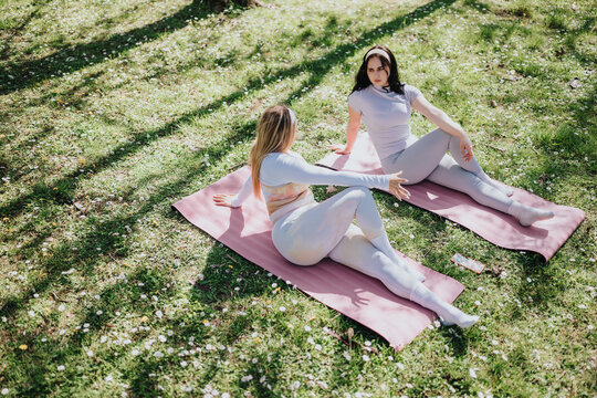 Two women practice seated yoga stretches on pink mats in a grassy park. The outdoor fitness session shows calm stretching and conversation during a sunny workout.