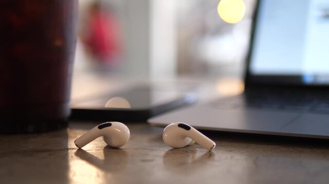 Cinematic close-up of white wireless earbuds on a cafe table with a blurred smartphone, laptop, and iced coffee in the background, modern remote workspace concept
