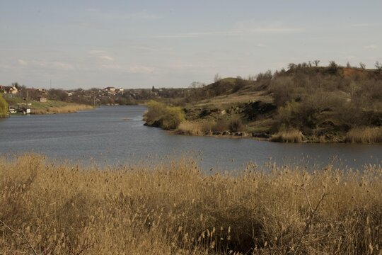 Calm river flowing through reeds and hills with rural houses in the background under a clear sky.