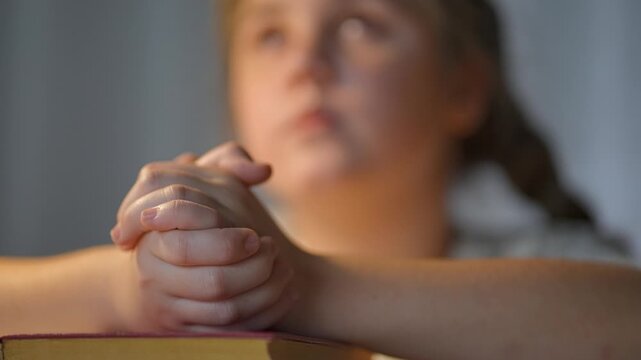 Girl praying at table with folded hands. Bible lies beneath praying hands. Child shows devotion in moment of quiet prayer. Girl closes eyes in trust. Faith shines in girl prayer and devotion.
