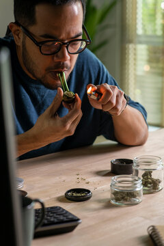 Man smoking a cannabis from smoking pipe, with cannabis equipment and accessories on the table.