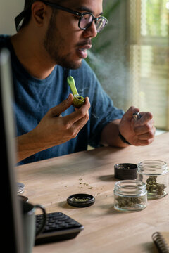 Man smoking a cannabis from smoking pipe, with cannabis equipment and accessories on the table.